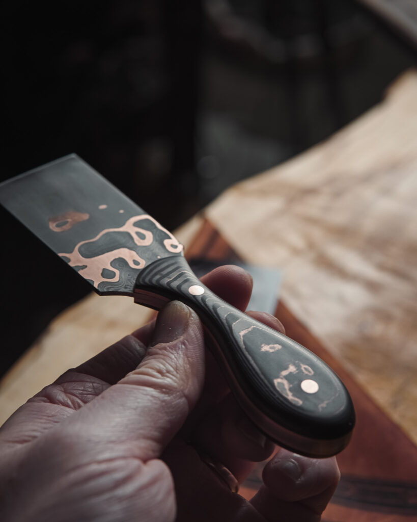 A hand holding a copper marbled-looking damascus steel skiving knife for leather work