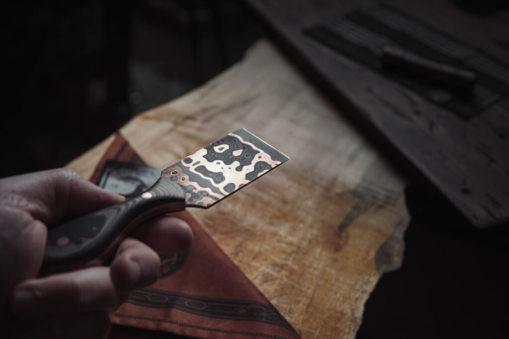 A hand holding a copper marbled-looking damascus steel skiving knife for leather work